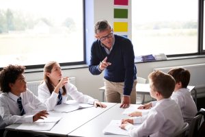 Male,High,School,Tutor,Standing,By,Table,With,Students,Teaching